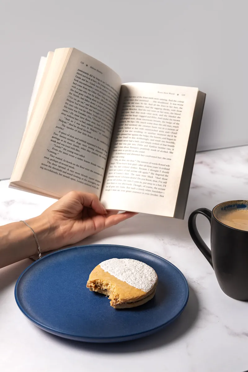 Concha mordida en plato azul junto a un libro abierto y una taza de café