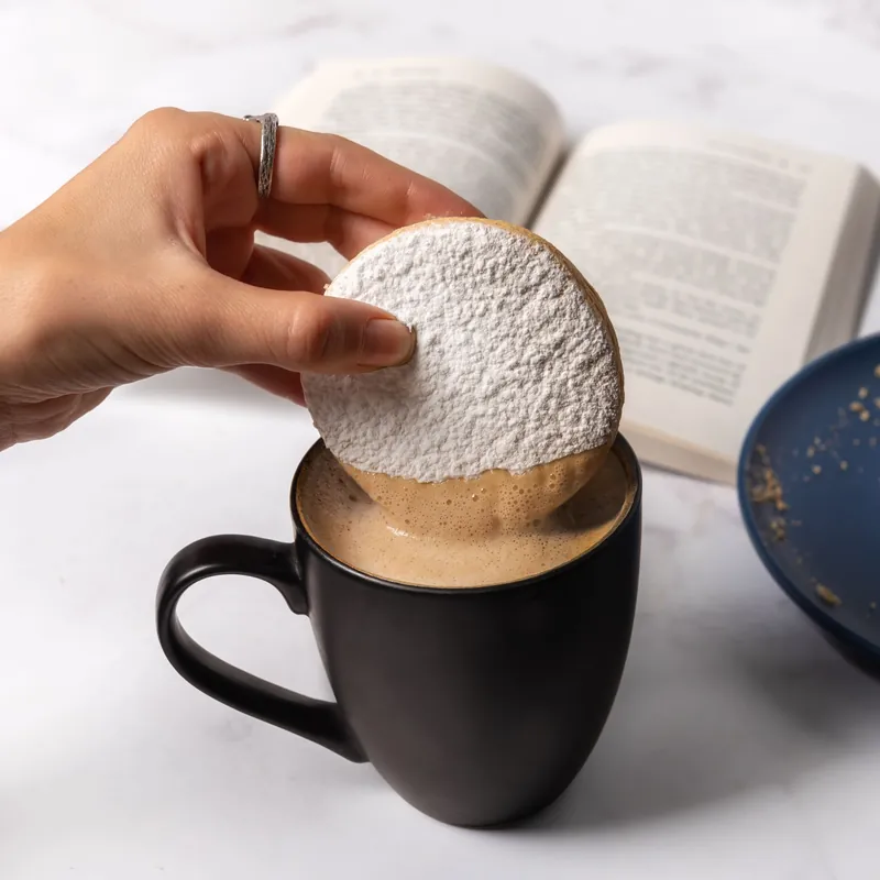 Concha espolvoreada con azúcar apoyada sobre una taza de café junto a un libro abierto