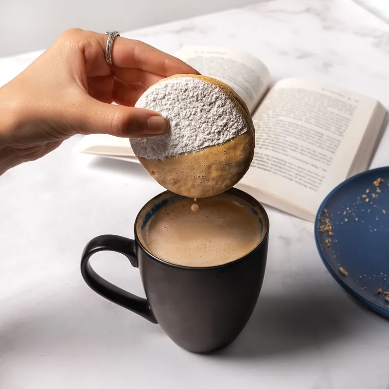 Mano colocando una concha con azúcar glass sobre una taza de café negro