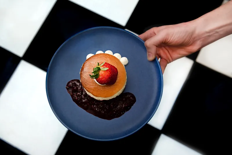 Mano sosteniendo un plato azul con cheesecake individual coronado con fresa sobre piso de ajedrez