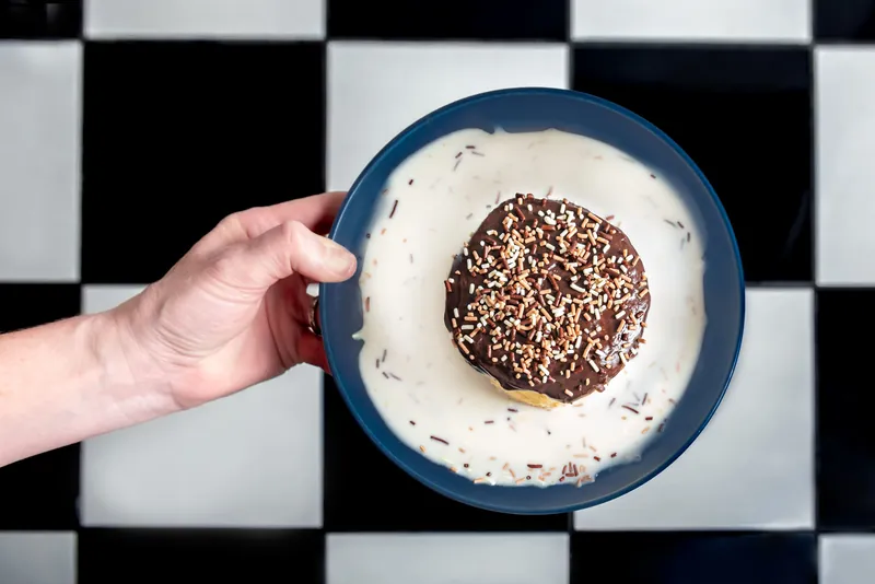 Mano sosteniendo un plato azul con dona de chocolate y nuez sobre piso de ajedrez